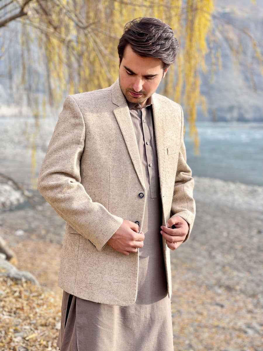 Man wearing a beige blazer and light-colored pants standing outdoors with trees and water in the background.