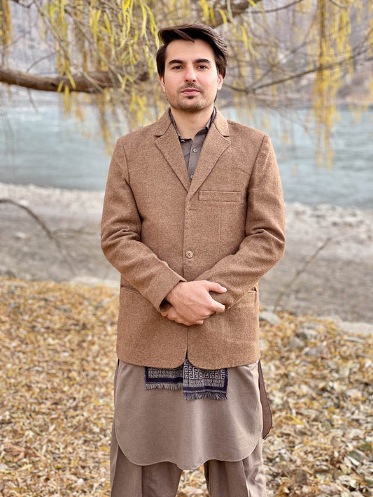 Man wearing a brown jacket and traditional outfit standing by a body of water with trees in the background.