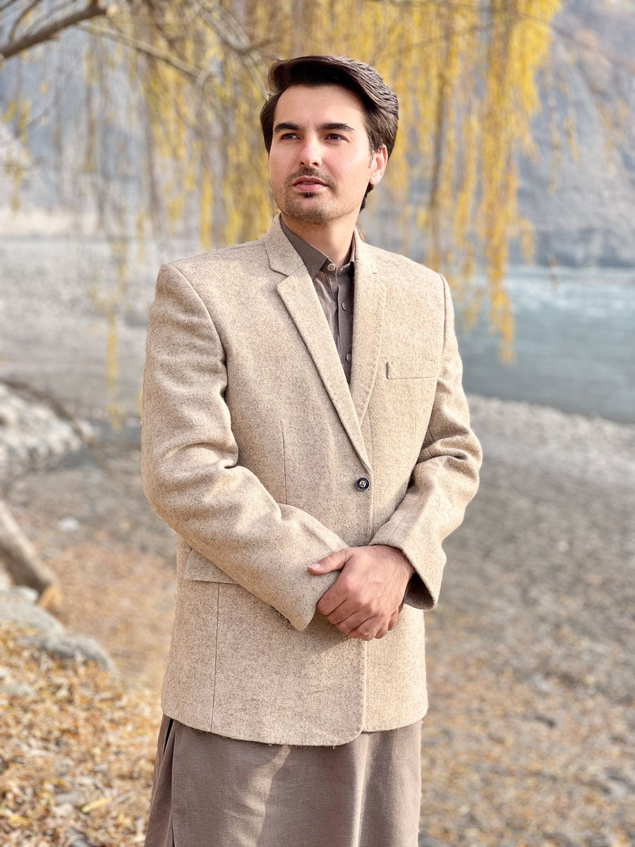 Man wearing a beige blazer standing outdoors with trees and water in the background