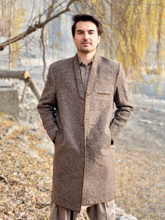 Man wearing a brown coat standing outdoors with trees and ground in the background